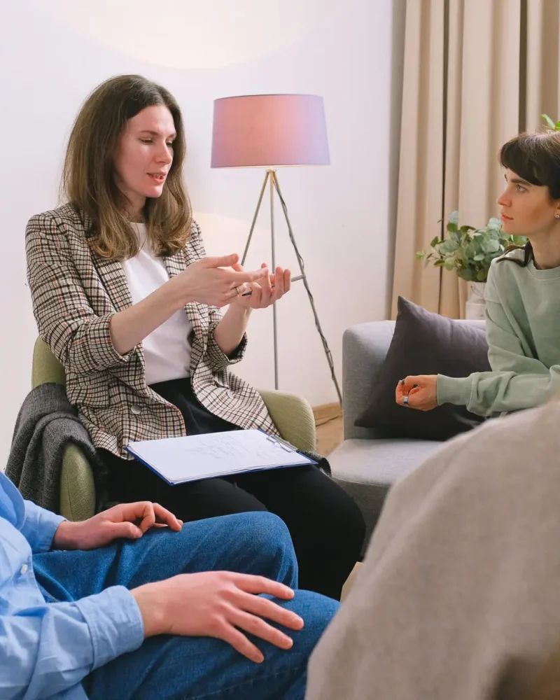 Young female therapist sitting on chair and discussing problem with patients during psychotherapy session in group